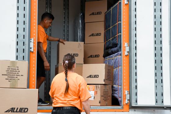 Two Allied movers in orange shirts are loading boxes labeled "Allied" into a moving truck